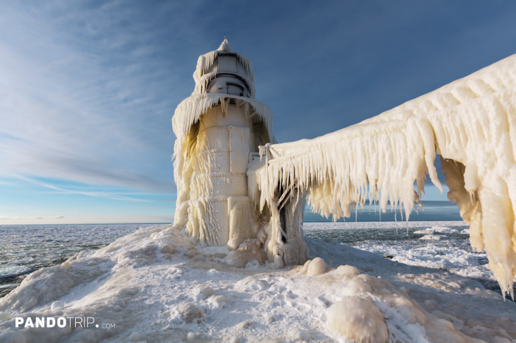Low-angle view of St. Joseph North Pier lighthouse encased in thick ice while its catwalk hangs with long icicles above a frozen Lake Michigan under clear winter light