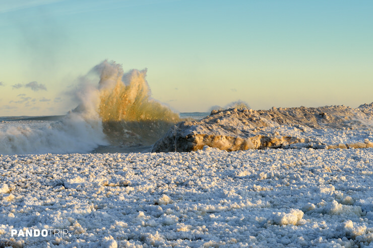Lake Michigan winter scene of pancake ice field and crashing wave against snow-covered ice ridge at sunrise, golden light on frozen shoreline