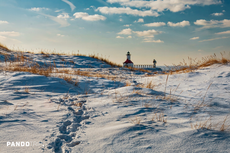 St. Joseph North Pier twin lighthouses framed by snow-covered Lake Michigan dunes, winter beach grass, blue sky, and sunlit footprints.