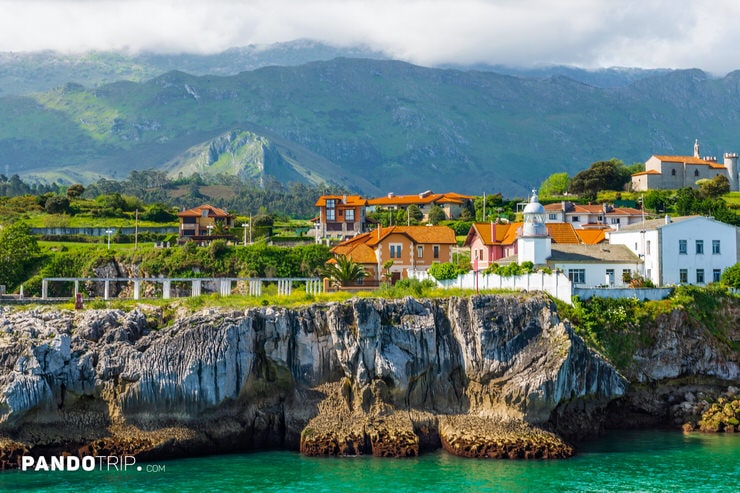 Seaside view of Ribadesella with orange-roofed houses, rugged cliffs, and green hills in the background.
