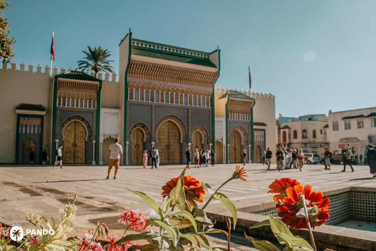 Tourists visiting the ornate golden gates of the Royal Palace in Fes el-Bali with red flowers in the foreground