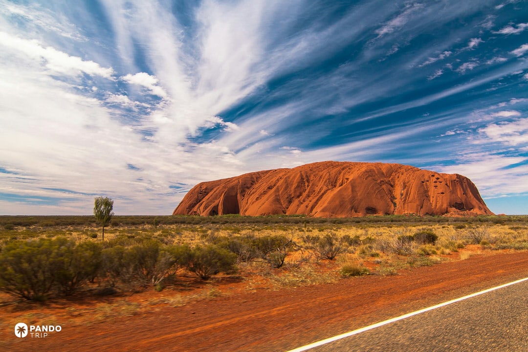 Daytime view of Uluru rising from flat desert scrub beside a red outback road in Uluru-Kata Tjuta National Park, Australia.