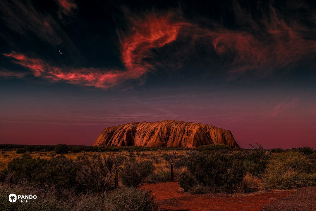 Uluru glowing deep red at dusk beneath dramatic clouds, framed by low desert shrubs in Uluru-Kata Tjuta National Park, Australia.