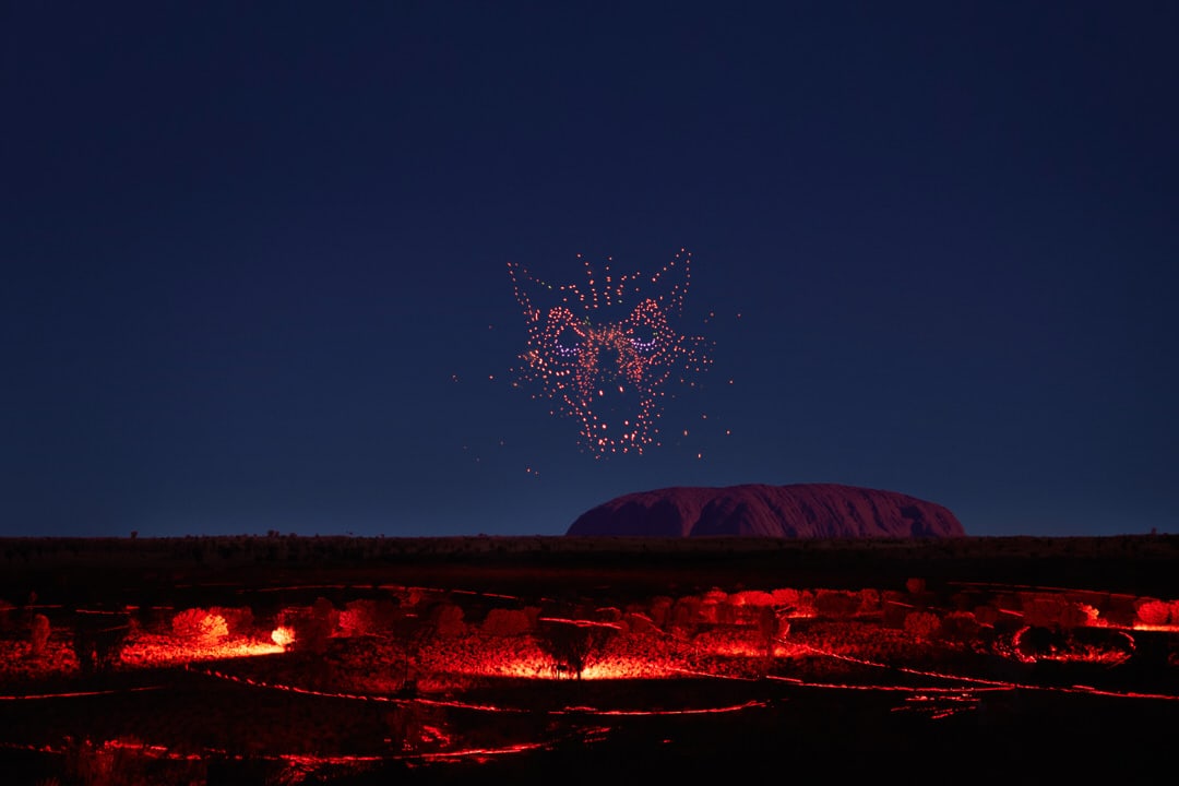 Wintjiri Wiru drone light show forming an Aṉangu story figure above Uluru at night, with the desert foreground glowing red in Uluru-Kata Tjuta National Park, Australia.