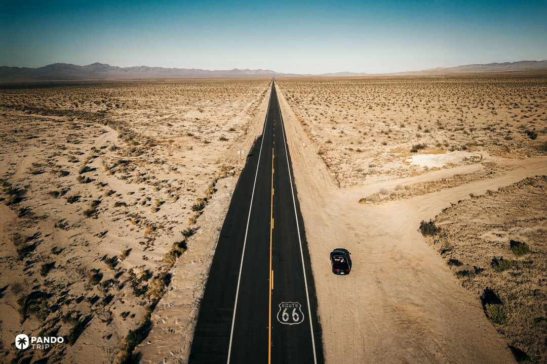 Aerial view of a straight two-lane Route 66 highway cutting through empty desert, with a single car parked on the shoulder in the American Southwest.