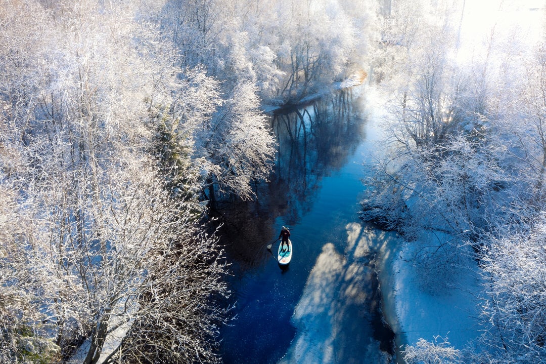A person stand-up paddleboarding on a narrow river lined with snow-covered trees on a bright winter day in Oulu, Finland.
