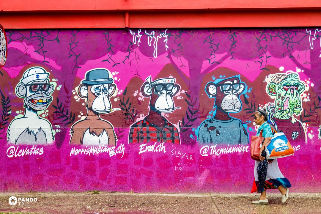 Street art mural in Comuna 13, Medellín, Colombia, with colourful cartoon ape characters on a bright pink wall and a local woman walking past.