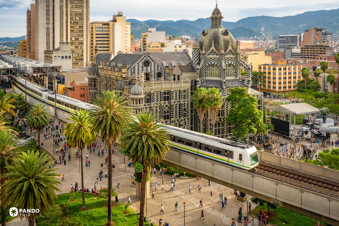 Medellín metro train passing the black-and-white Rafael Uribe Uribe Palace of Culture and a palm-filled plaza in the city centre.