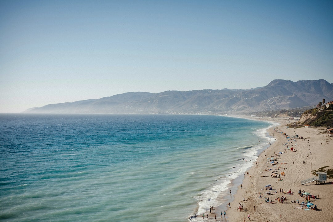 Wide landscape view of Zuma Beach in Malibu showing white sand, blue ocean, and Santa Monica Mountains in the background.