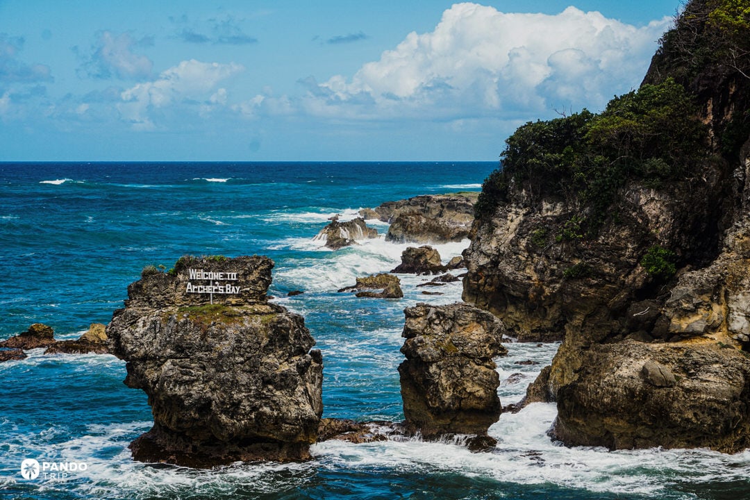 Rocky cliffs and crashing Atlantic waves at Archer’s Bay on the rugged north coast of Barbados, with a “Welcome to Archer’s Bay” sign on a sea stack.