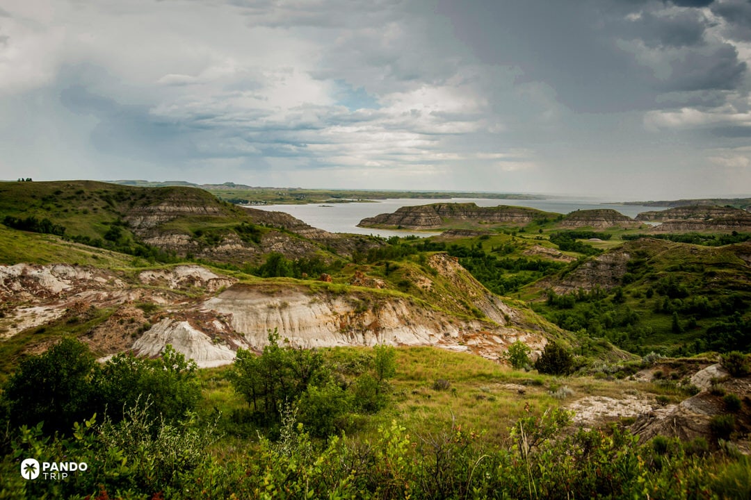 View over Lake Sakakawea in western North Dakota, with layered badlands hills, green valleys and rocky shoreline under a cloudy sky.