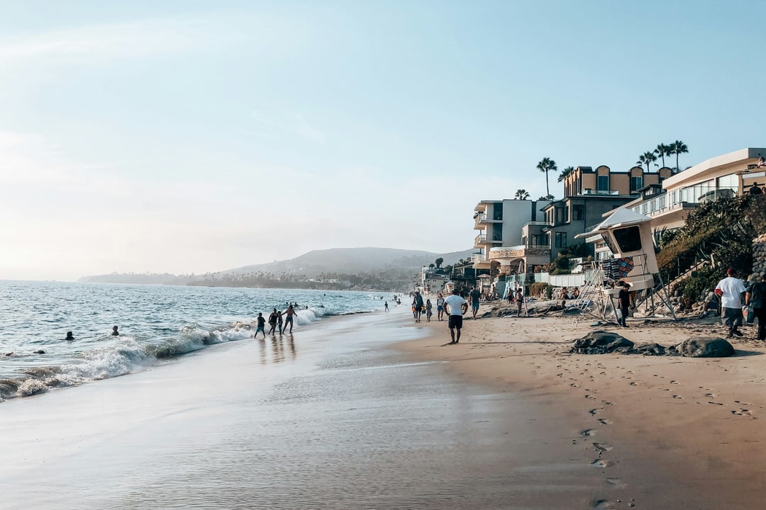 People walking on the sand at Sleepy Hollow Beach in Laguna Beach with beachfront houses and a lifeguard tower.