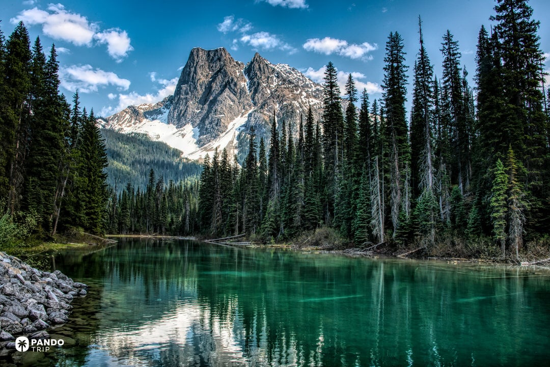 Turquoise alpine lake ringed by dense evergreen forest with a steep snow-dusted peak rising behind in Columbia-Shuswap A, British Columbia, Canada.