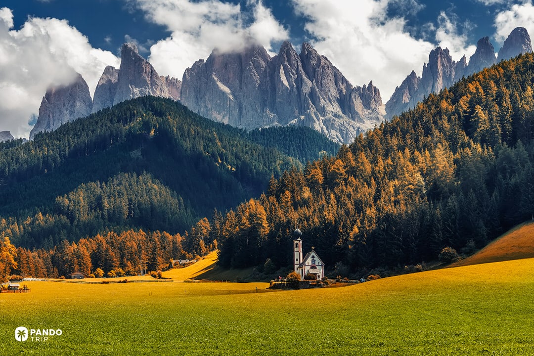The Church of St. Johann in Val di Funes surrounded by yellow larch trees and the Odle peaks in the Dolomites.