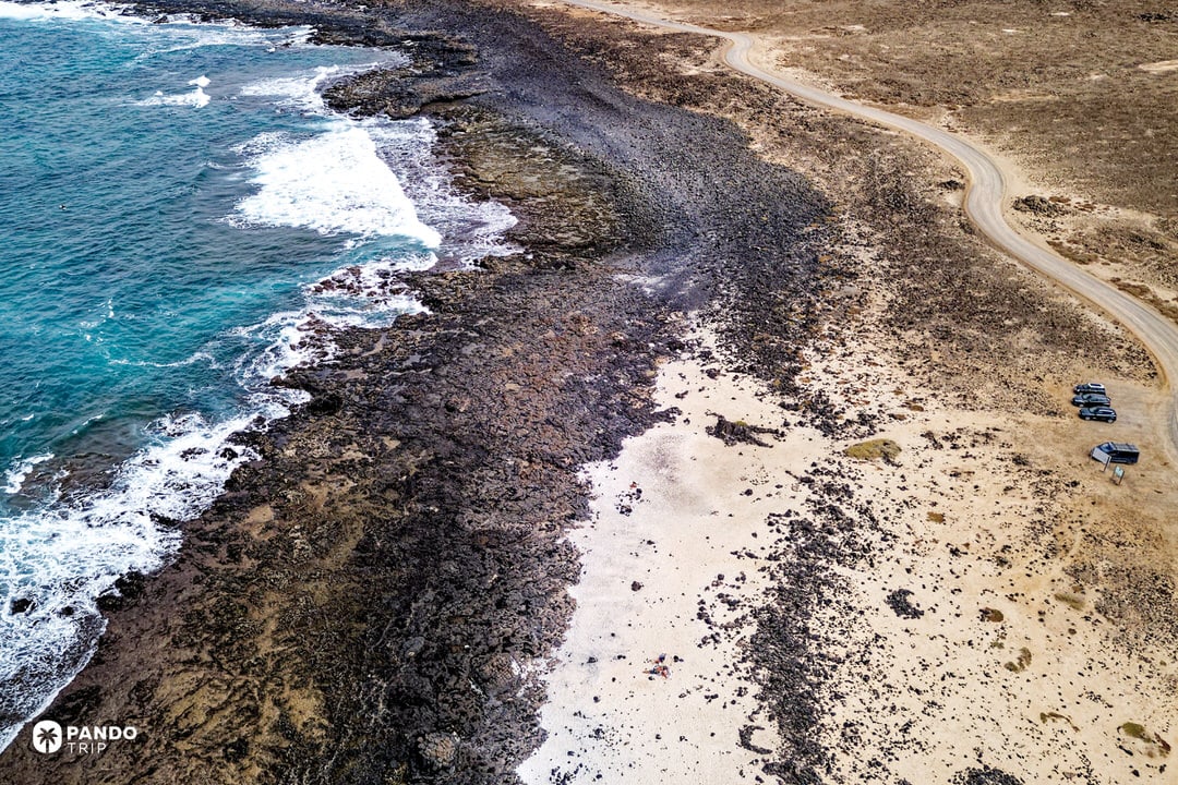 Aerial of Popcorn Beach where black lava flows meet a pale rhodolith shore and dirt access track, Fuerteventura.