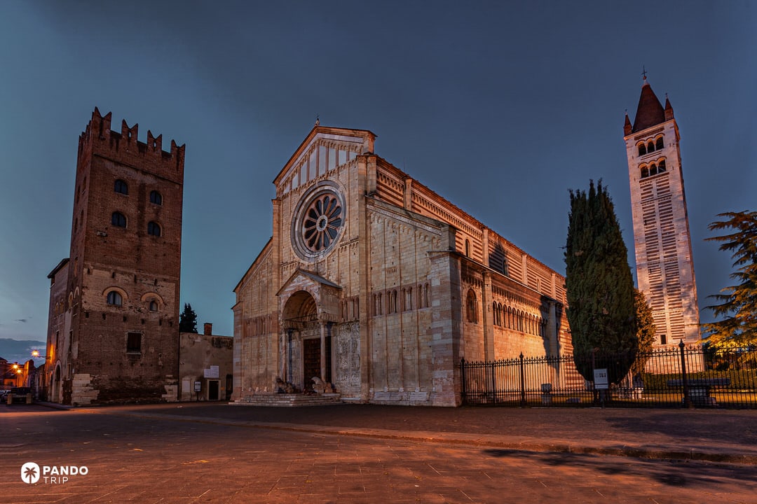 The illuminated facade and bell tower of Basilica di San Zeno Maggiore in Verona against a dark blue twilight sky.