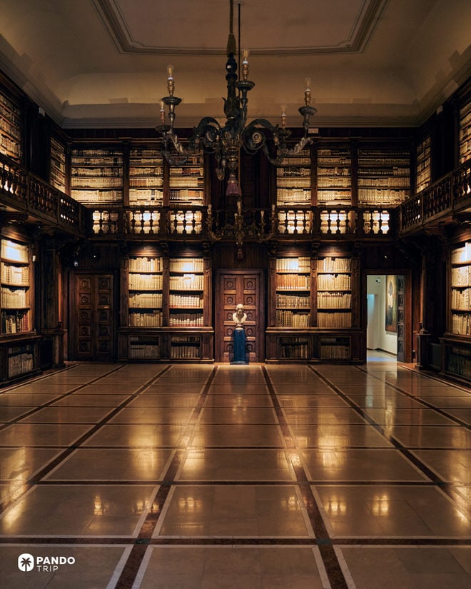 Interior of the Biblioteca Capitolare in Verona showing ancient wooden bookshelves, a tiled floor, and a chandelier.