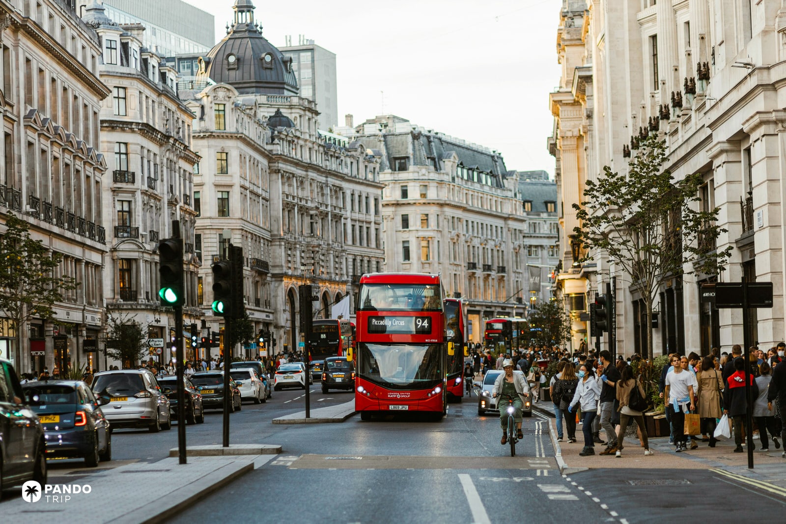 A busy London street scene looking down Regent Street, featuring a bright red double-decker bus headed to Piccadilly Circus, surrounded by heavy traffic, crowds of pedestrians, and classic stone architecture.