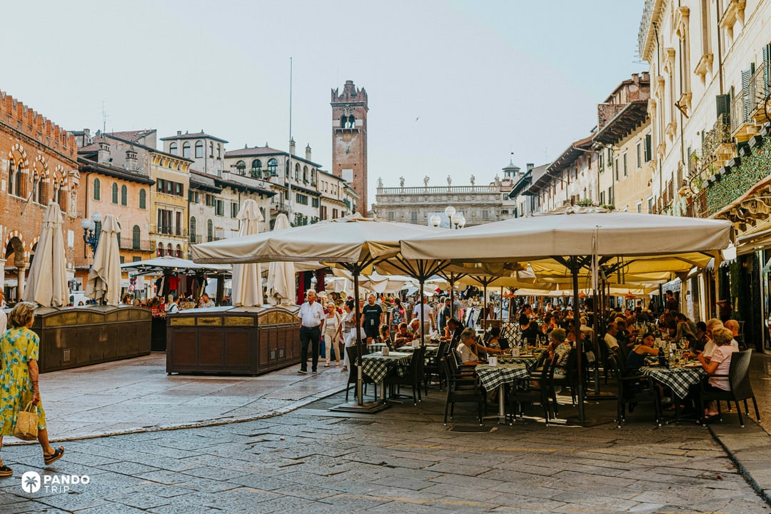 Ground level view of Piazza delle Erbe in Verona during the day, crowded with white umbrellas, restaurant tables, and tourists walking across the stone pavement.