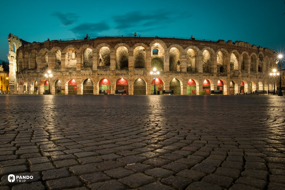 The exterior arches of the Verona Arena illuminated at night, viewed from the cobblestone pavement of Piazza Bra against a dark teal sky.