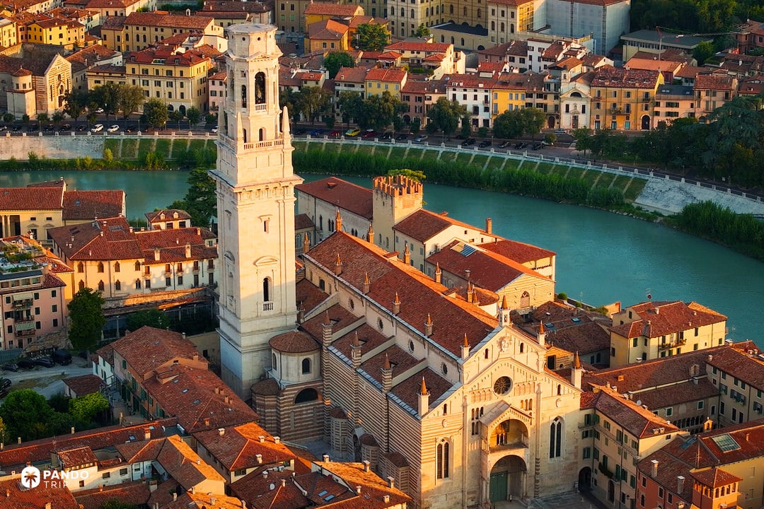 Close-up aerial view of the Verona Cathedral and bell tower glowing in warm golden hour sunlight next to the teal Adige River.