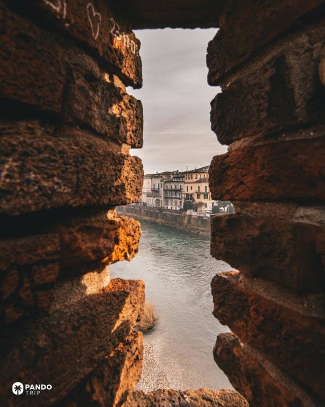 View of the Adige River and Verona buildings framed through a brick arrow slit in a fortified wall.
