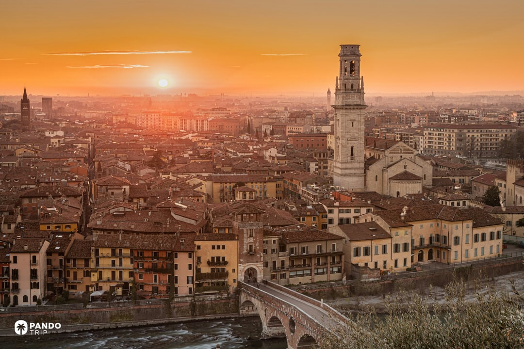 Sunset view from Castel San Pietro overlooking the Ponte Pietra bridge, the Adige River, and the Verona skyline bathed in orange light.