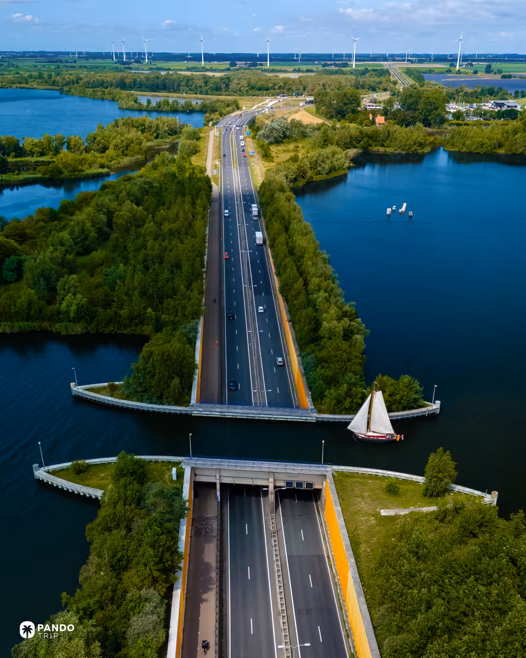 The N302 running north from Harderwijk into the Aquaduct Veluwemeer, a traditional Dutch sailing barge crossing the water above the tunnel mouth, wind turbines on the Flevoland horizon.