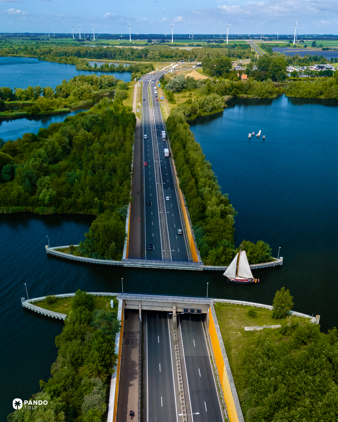 The N302 running north from Harderwijk into the Aquaduct Veluwemeer, a traditional Dutch sailing barge crossing the water above the tunnel mouth, wind turbines on the Flevoland horizon.