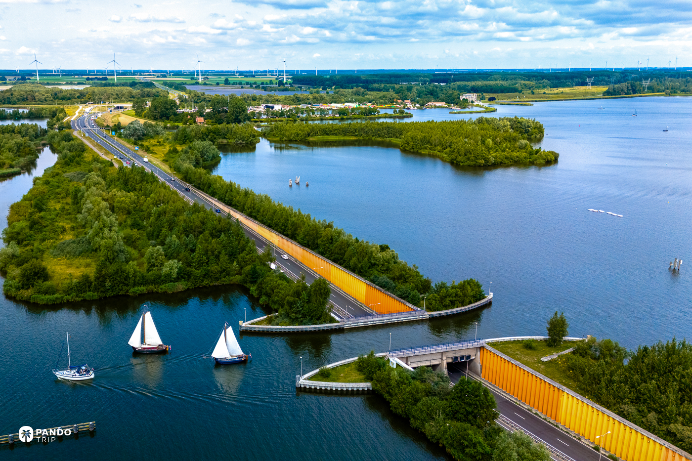 Wide aerial view of the Aquaduct Veluwemeer near Harderwijk, three sailing boats waiting on the lake as the N302 drops into the tunnel beneath the water.