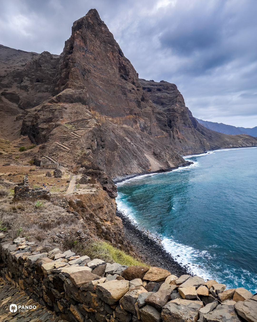 A dark volcanic peak above terraced slopes and a black pebble beach on the coast at Aranhas, Santo Antao, Cape Verde.