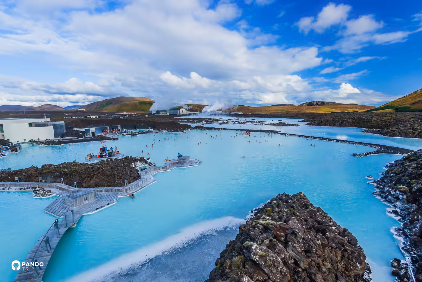 Bathers in the pale blue water of Iceland's Blue Lagoon, edged by black lava and low buildings.