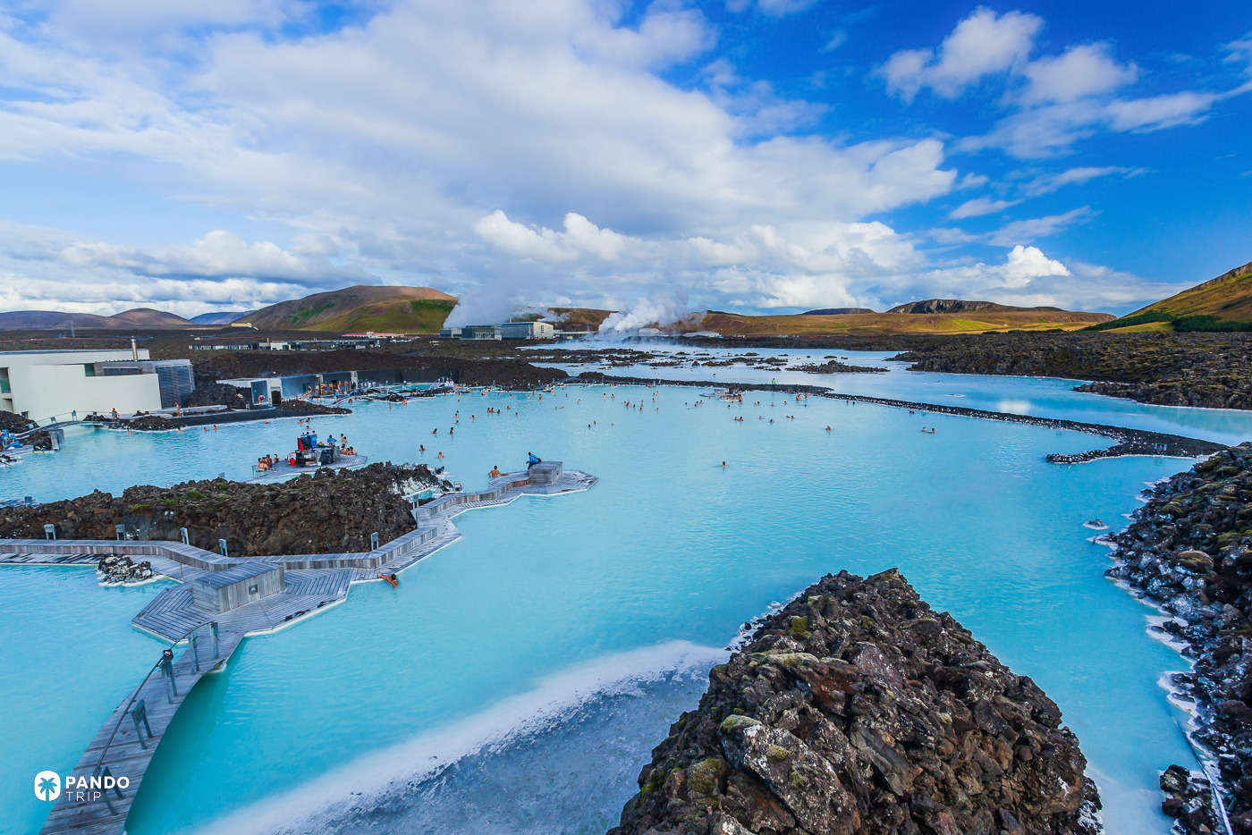Bathers in the pale blue water of Iceland's Blue Lagoon, edged by black lava and low buildings.