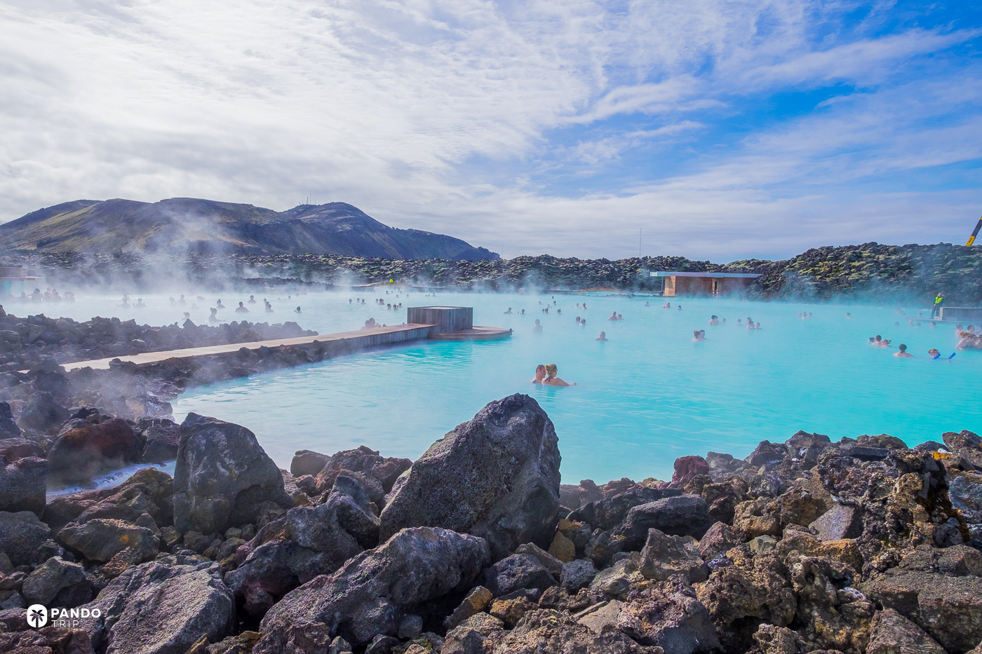 Steam over the Blue Lagoon, with lava boulders in the foreground and bathers in the milky water.
