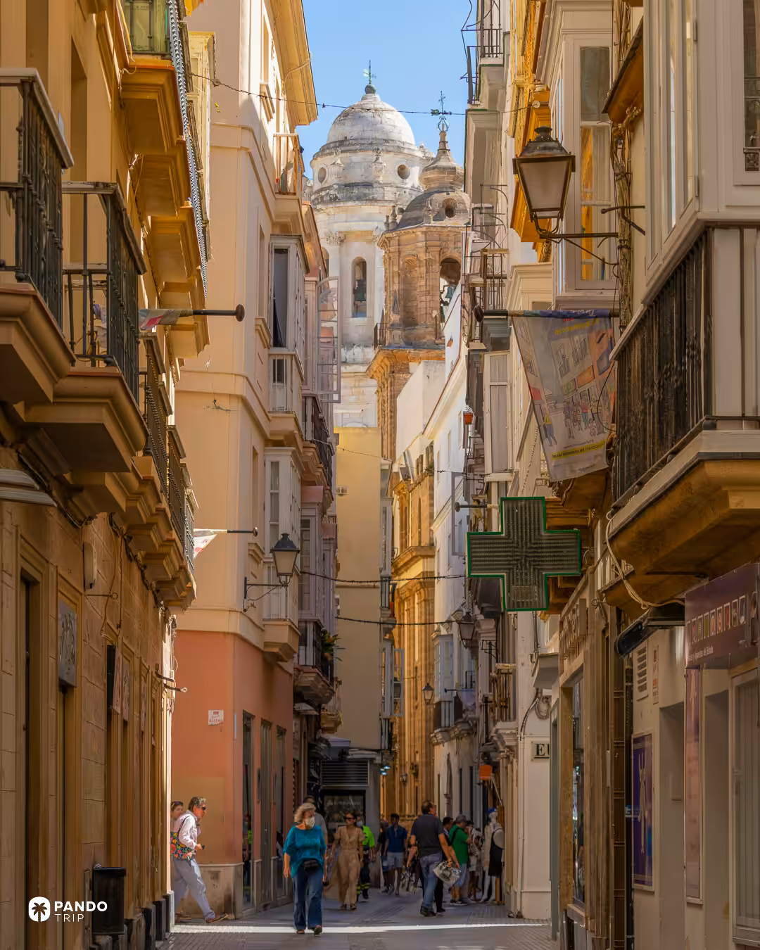 People walking a narrow street in Cadiz between bay-windowed houses, the cathedral's white dome and belfry framed at the end of it.