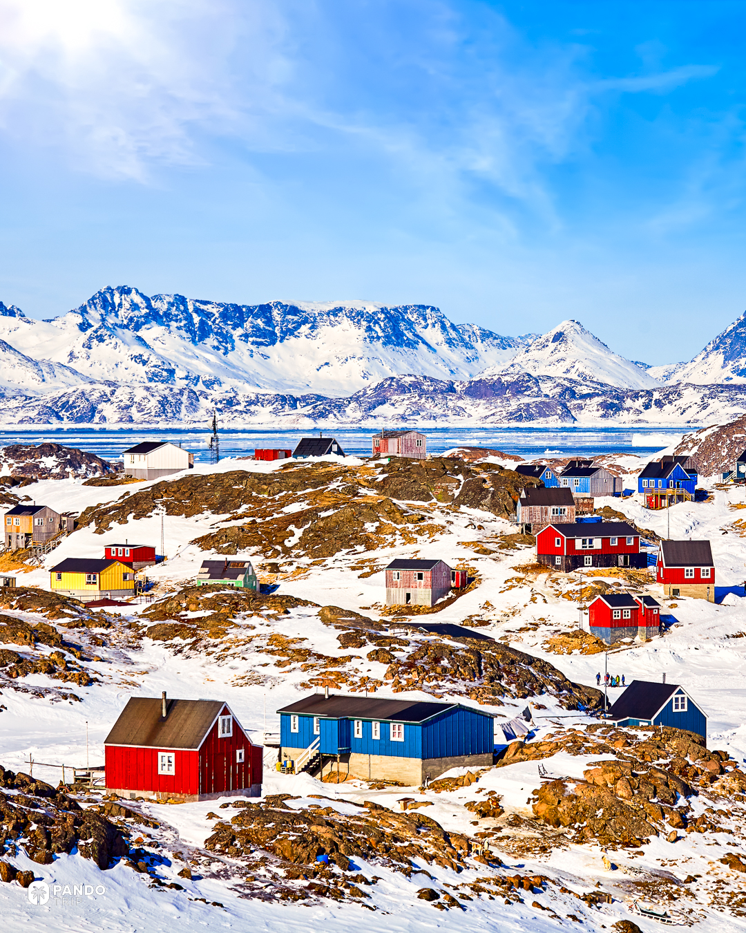 Red, blue and yellow timber houses on snow-covered rock above sea ice at Kulusuk, east Greenland, with snow peaks behind.