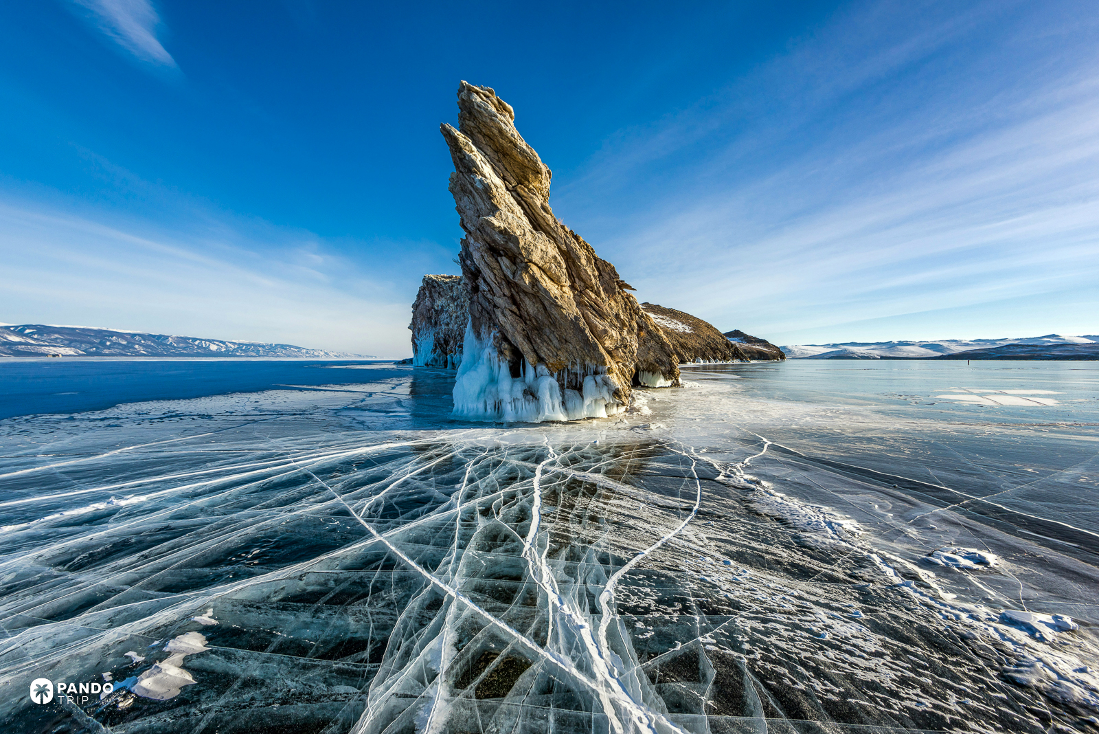 Cracked transparent ice on frozen Lake Baikal with an ice-fringed rock outcrop behind.