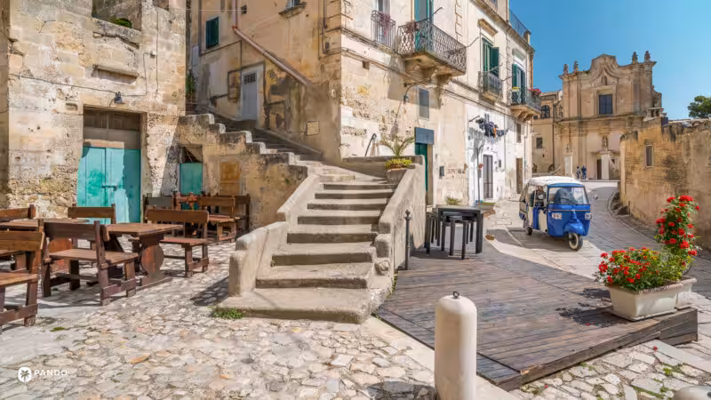 A blue three-wheeled Piaggio Ape on the cobbles of a square in Matera's Sassi, cafe benches and a baroque church facade behind it.