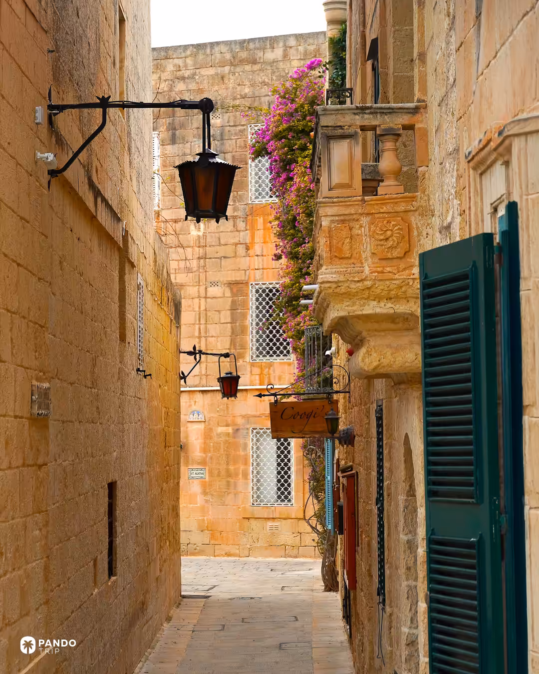 A narrow honey-coloured limestone lane in Mdina with a wrought-iron lantern and bougainvillea above a green-shuttered doorway.