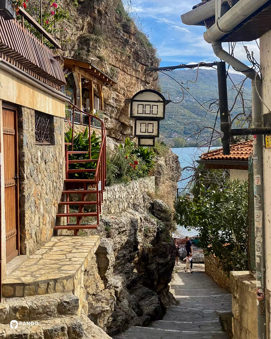 A stepped lane in Ohrid running down to the lake beneath a rock face, a red spiral staircase and a lantern above it.