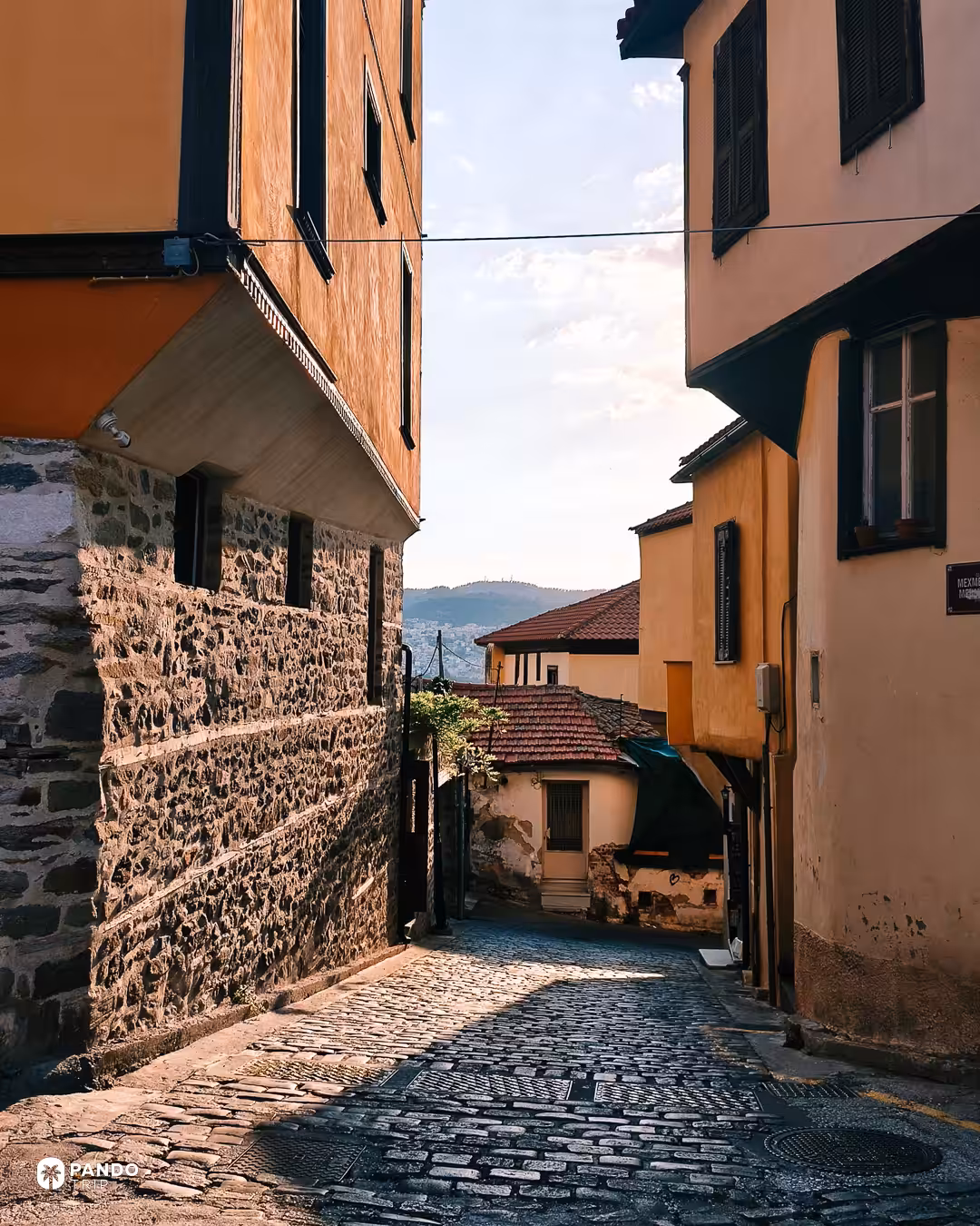 A steep cobbled lane between overhanging Revival houses in Plovdiv, the hills beyond the city visible at the end of it.