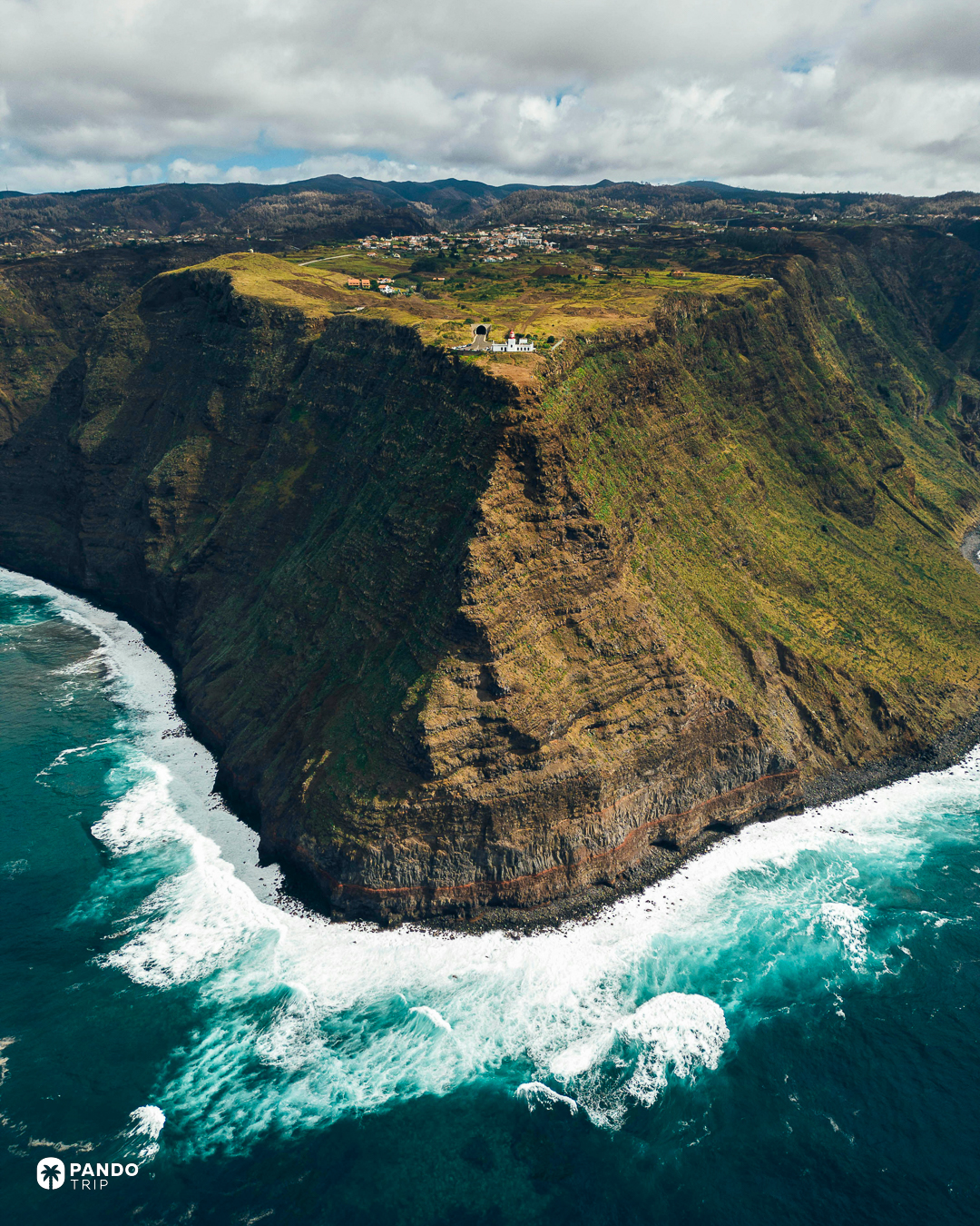A white lighthouse on the cliff edge at Ponta do Pargo with farmland behind it and surf breaking on the rocks far below, Madeira.