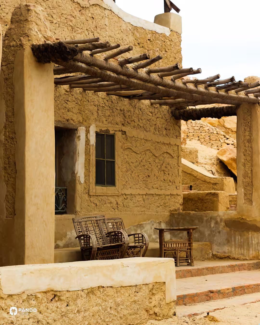 A Siwan building of salt-mud kershif with a palm-log pergola and carved wall relief, rattan chairs on the terrace beneath it.