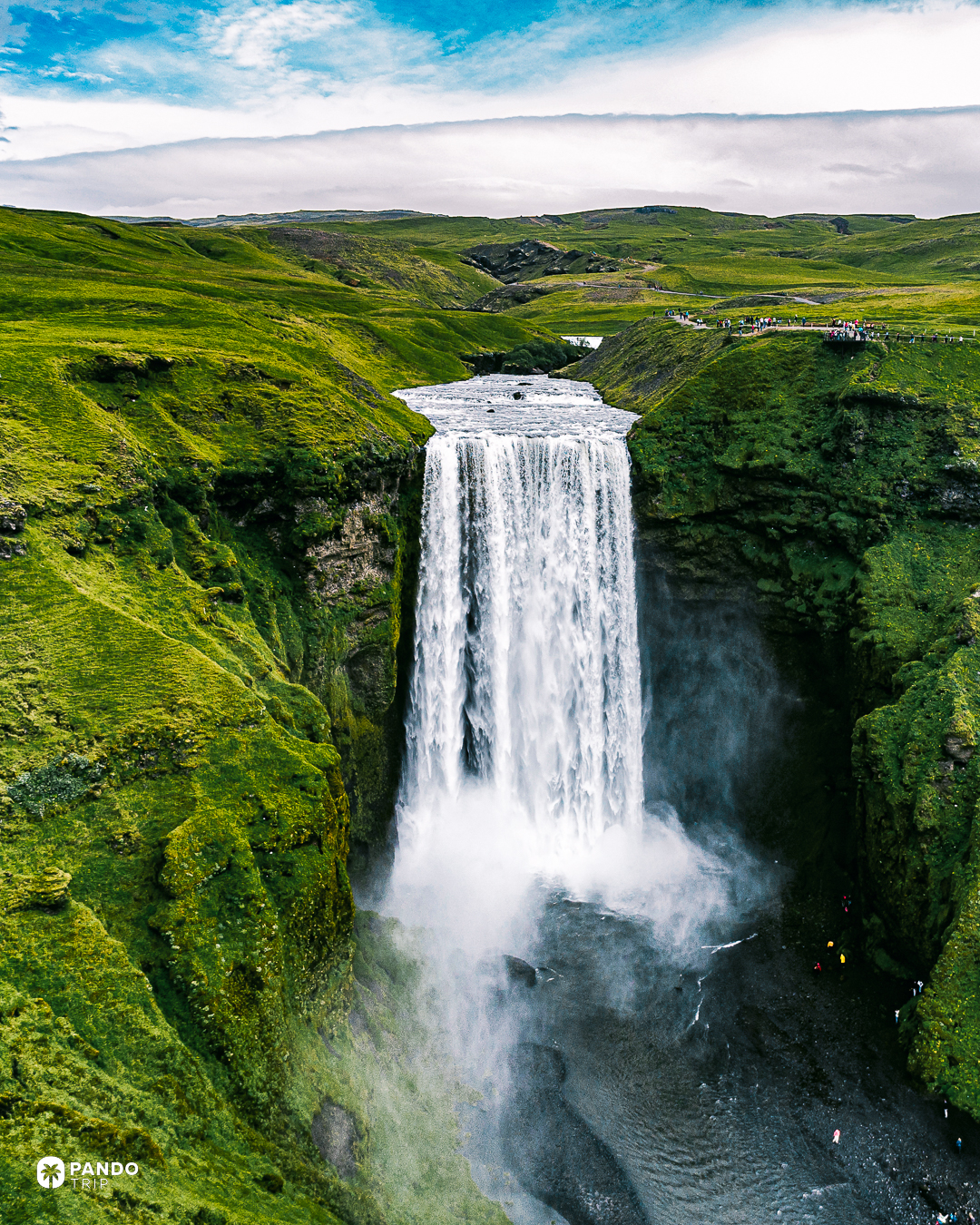 The Skogafoss waterfall dropping between green cliffs in southern Iceland, with visitors on the path along the top.