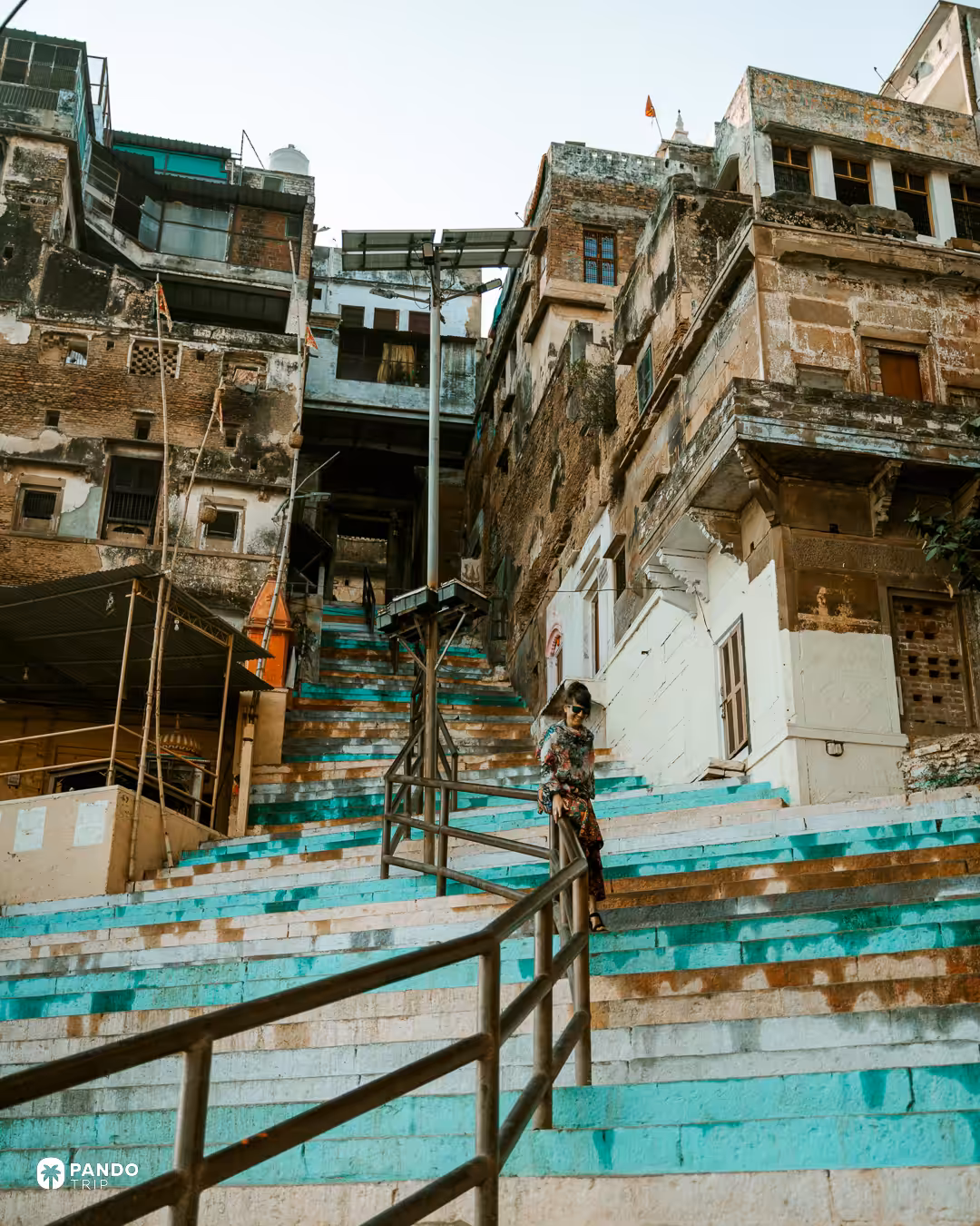 Turquoise-painted ghat steps in Varanasi climbing to weathered riverfront houses, a woman standing halfway up them.