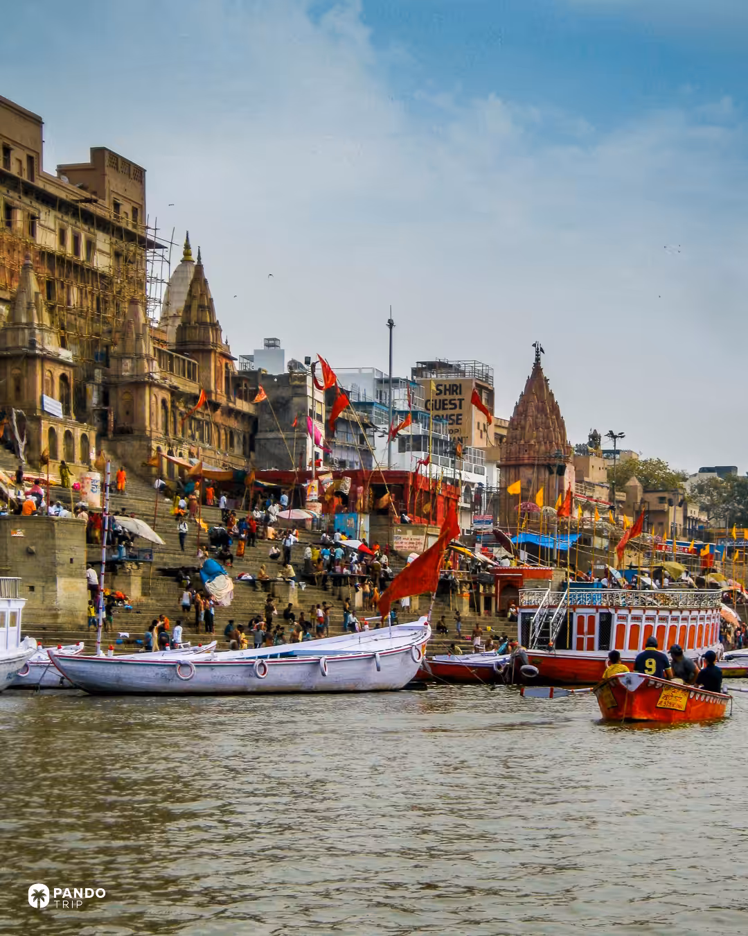 Temple spires and crowded steps on Varanasi's riverfront seen from the water, red and yellow flags along the ghat and boats moored below.