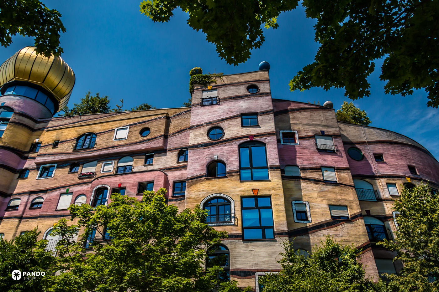 Banded ochre and pink walls of the Waldspirale, no two windows alike, with trees growing along the roofline.