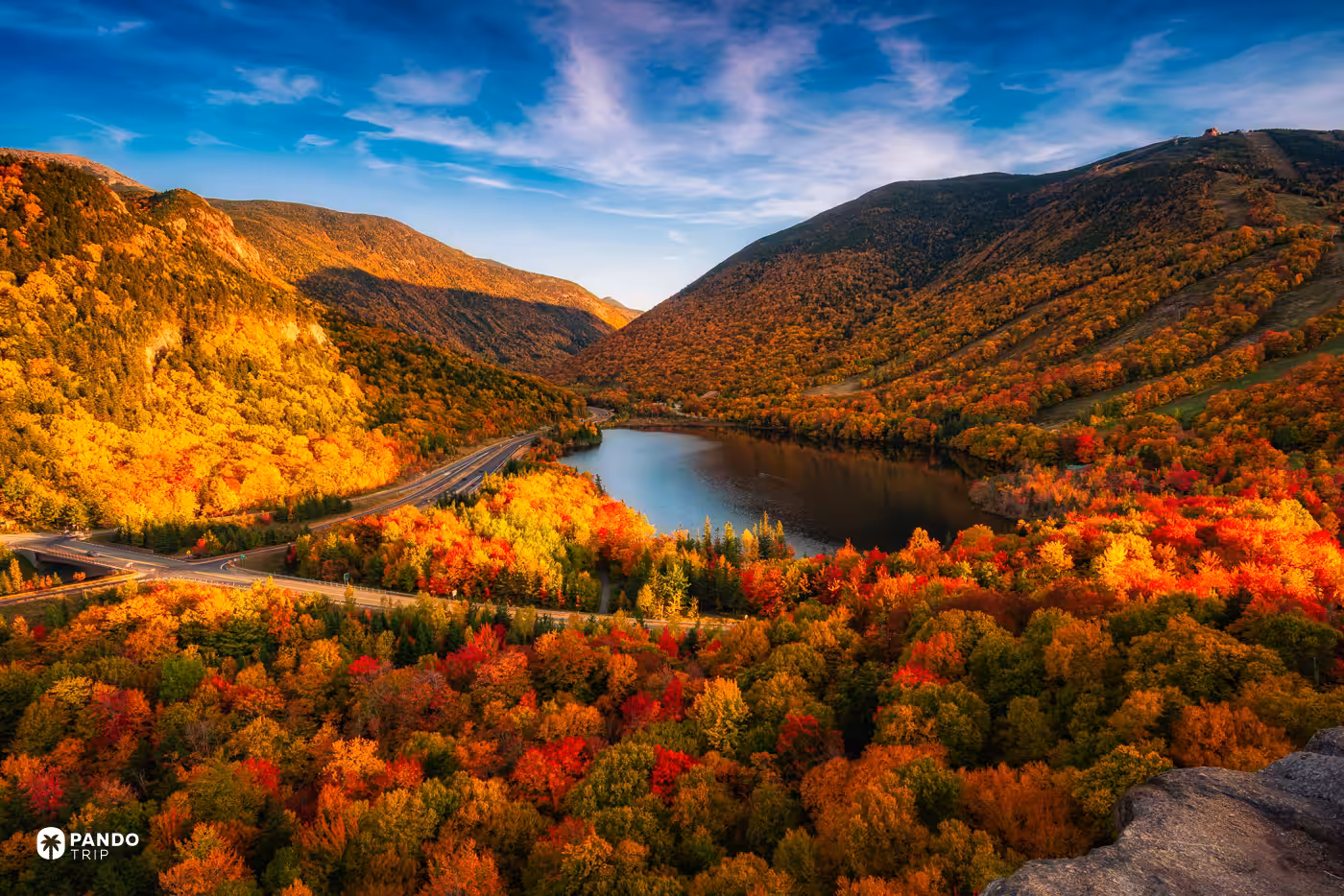 Echo Lake below Cannon Mountain in Franconia Notch, New Hampshire, surrounded by red, orange and yellow fall foliage under a blue sky.