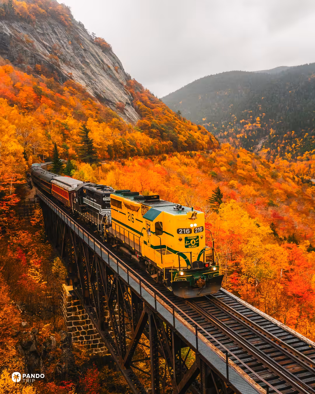 A Conway Scenic Railroad train on the Frankenstein Trestle beneath a granite cliff and bright autumn forest in Crawford Notch.
