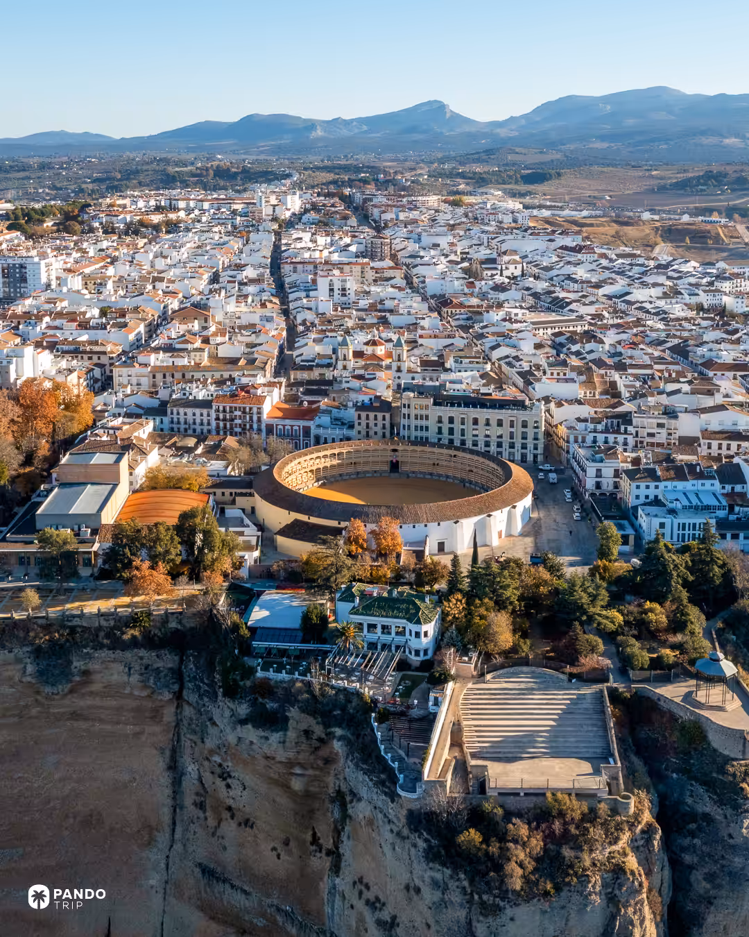 Aerial view of Ronda's round bullring and the grid of white streets behind it, with the plateau's cliffs in the foreground and mountains on the horizon.